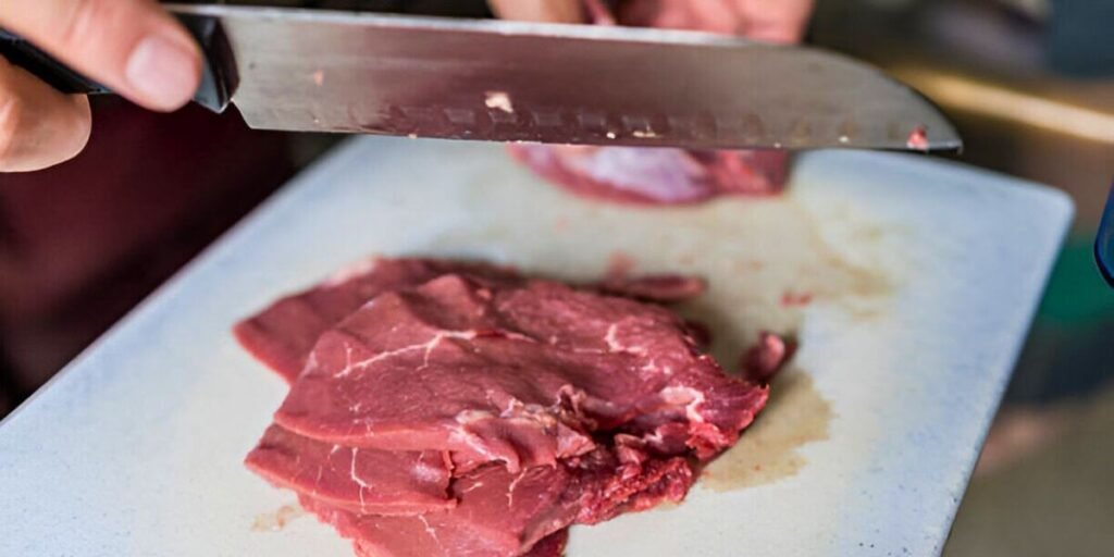 Close-up of hands using a Santoku knife to slice meat on a cutting board