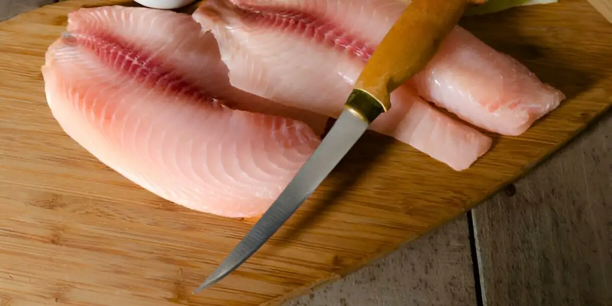 A fillet knife with a wooden handle placed on a cutting board with fresh fish fillets.